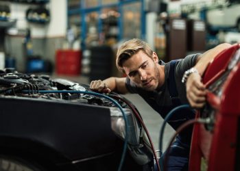 Young auto mechanic using compressor while maintaining AC unit of a car at workshop.